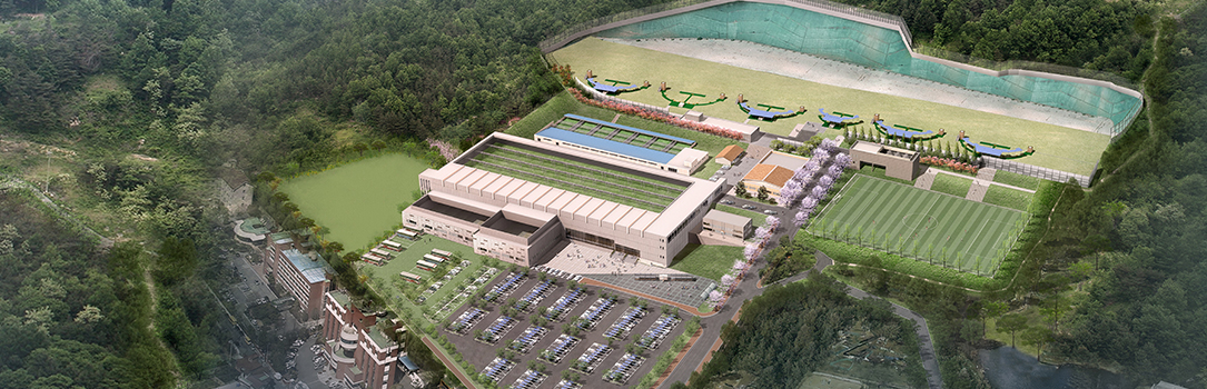 Aerial view of a large sports complex with buildings, a parking lot, and fields surrounded by dense green forest on a calm day